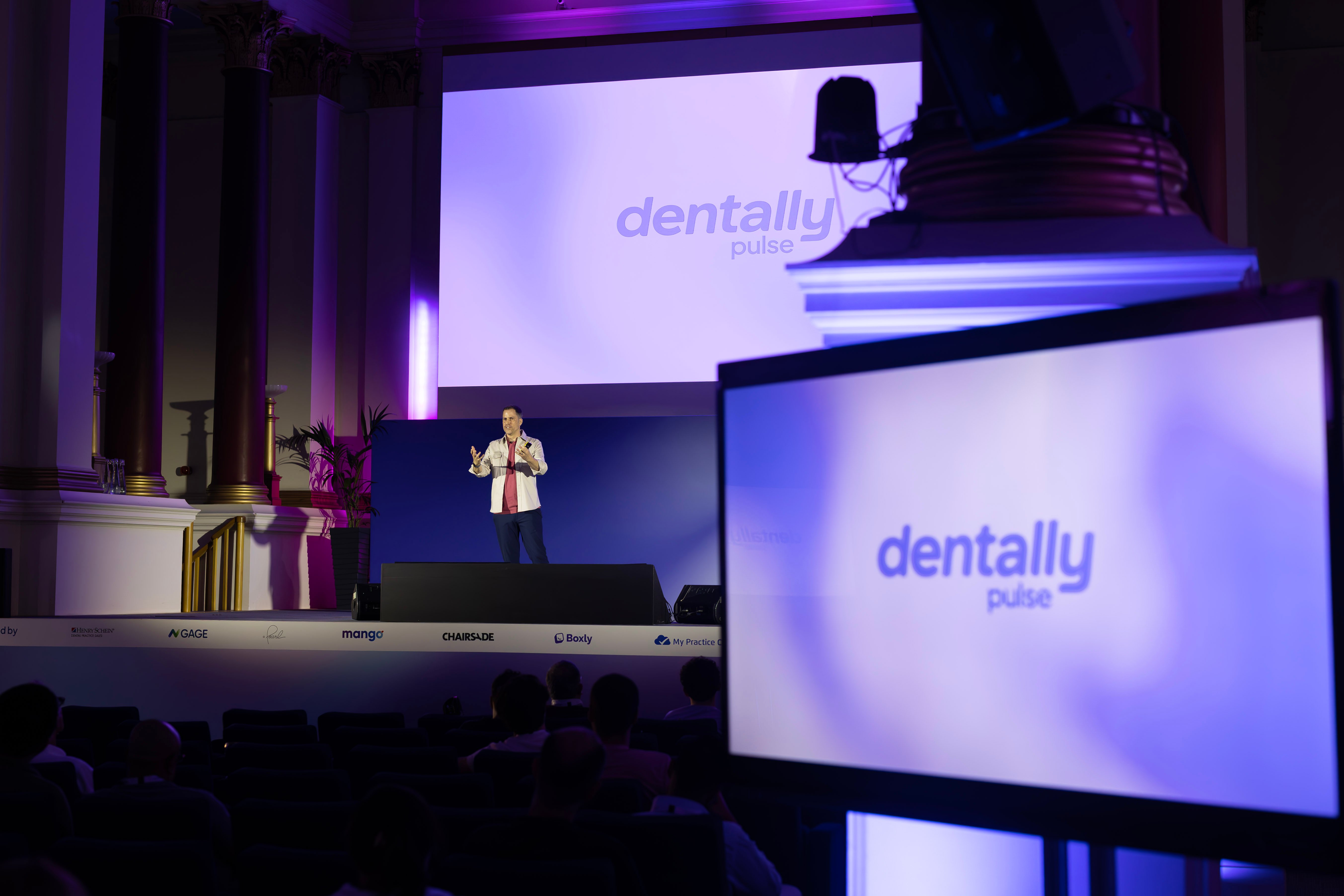 A speaker presenting Dentally Pulse on stage to an audience, with the Dentally Pulse logo on the screens behind