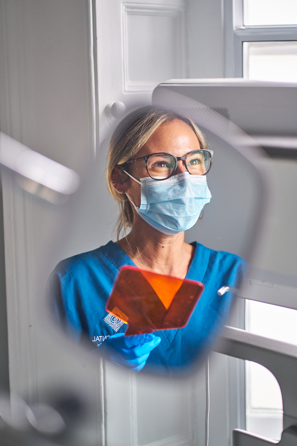 A photograph of a dental mirror with a dental nurse wearing glasses and a mask in the mirror